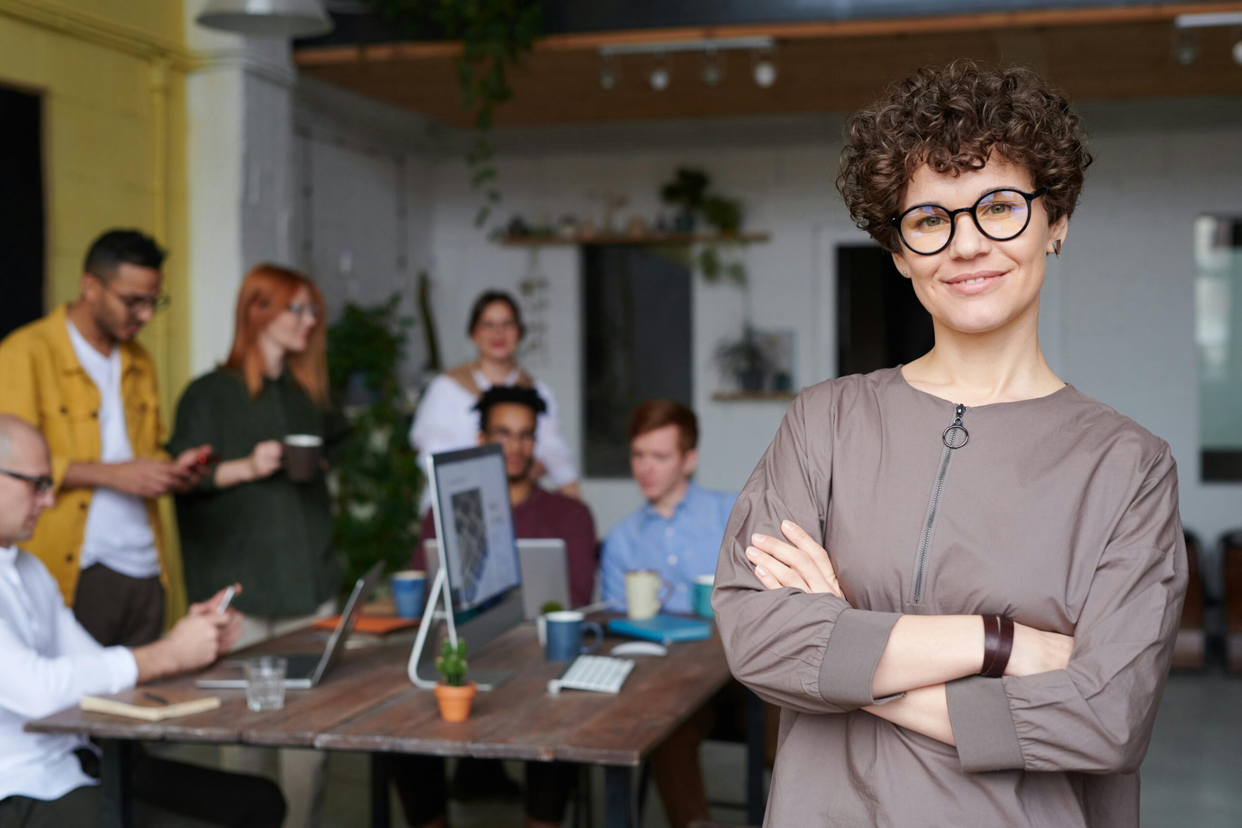 Home Smiling businesswoman with curly hair stands confidently in a modern office space with colleagues.